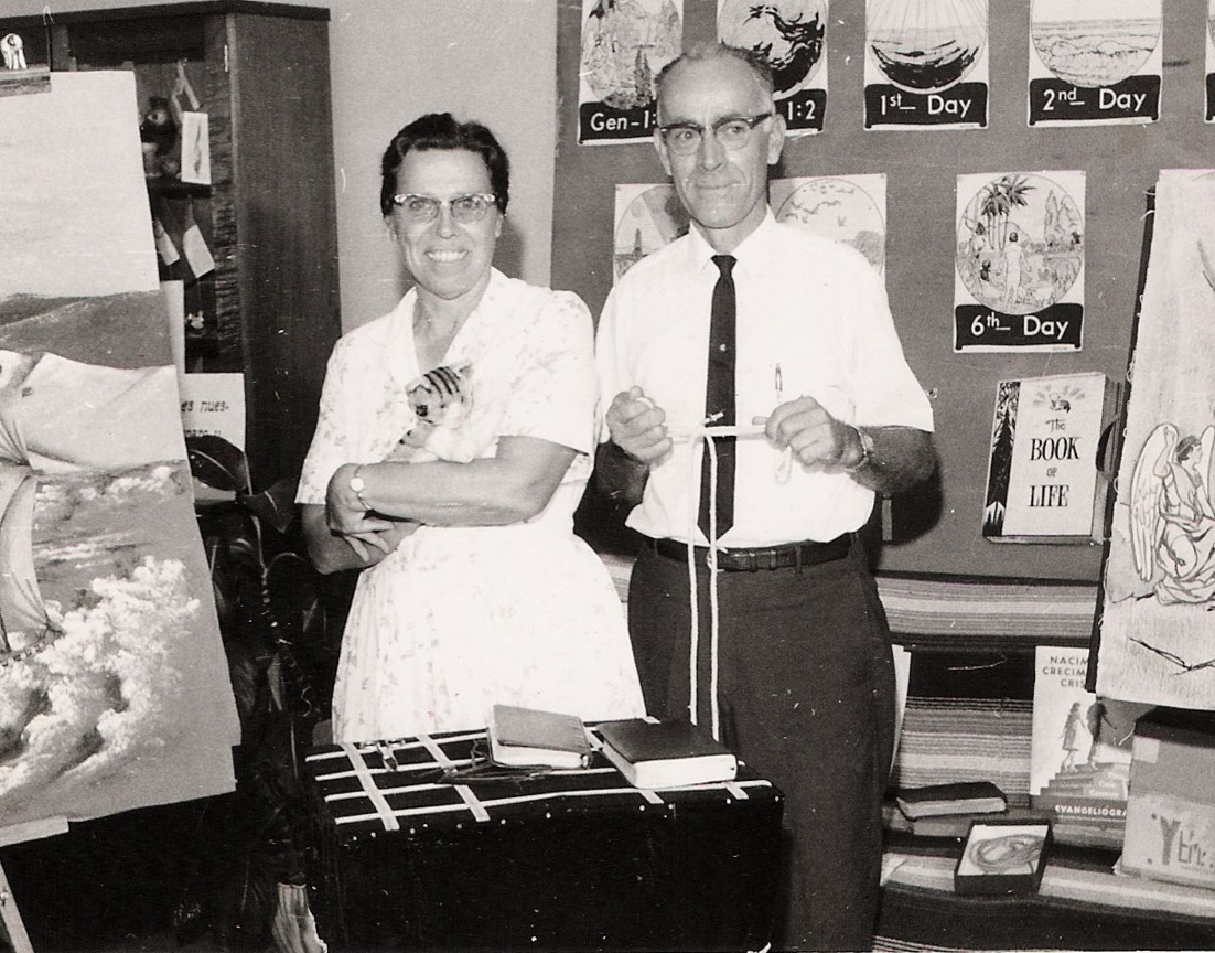 Pearl and Howard Wolgemuth stand in the center of a small display area. Pearl is holding a hand puppet shaped like a cat. Behind them are various images of Bible scenes as well as a flannelgraph board.