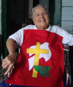 A woman sits in a chair with a hand-sewn version of the Brethren in Christ logo on her lap. The logo is a yellow cross on top of a white dove and a basin with a towel.